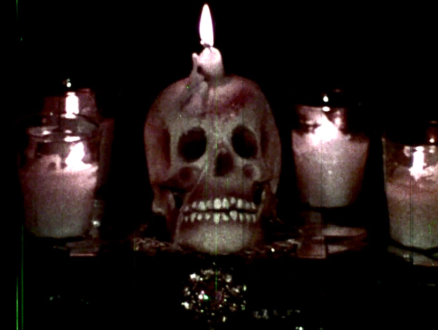 A skull with a candle on top of it sits center of frame in a darkened room, between more candles. A skull with a candle on top of it sits center of frame in a darkened room, between more candles.