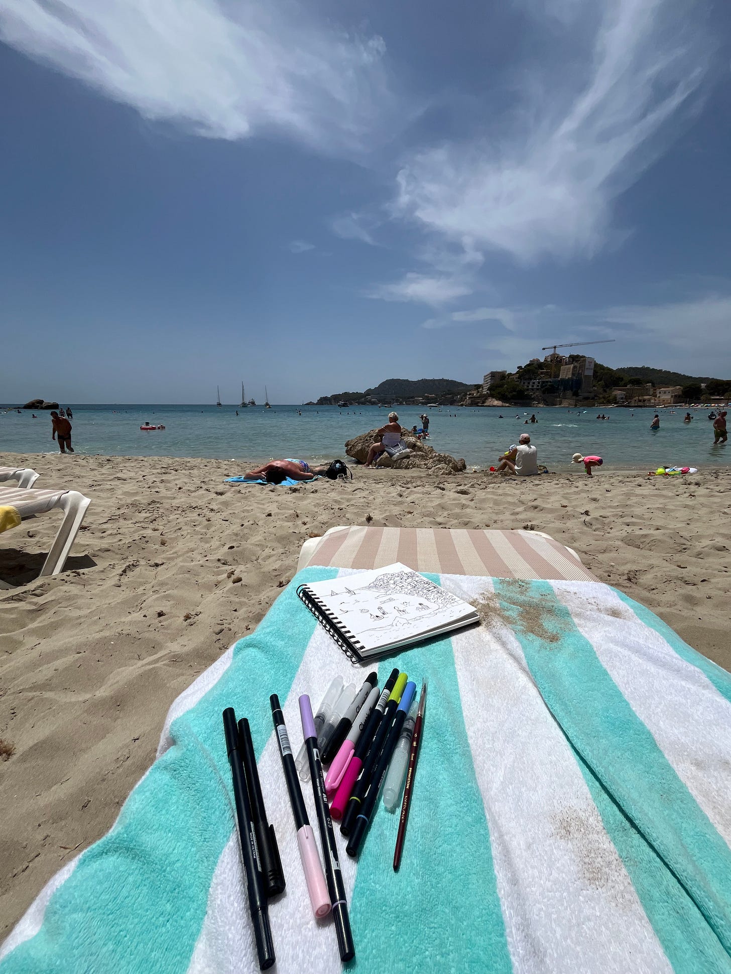 My view from my beach chair. Looking out at the ocean, laying on a blue and white striped towel, markers and pens and watercolors sitting next to the sketch of what will become the "Topless in Mallorca" piece