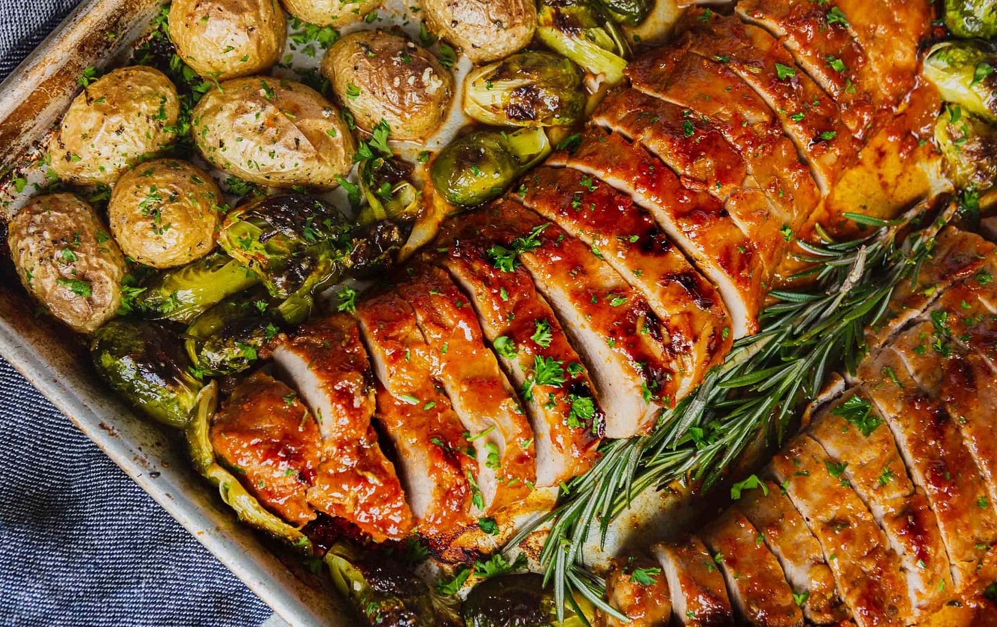 A sheet pan with two glazed, roasted pork loins. There are potatoes and brussels sprouts on the sheet pan also. The pork loins are sliced for serving.