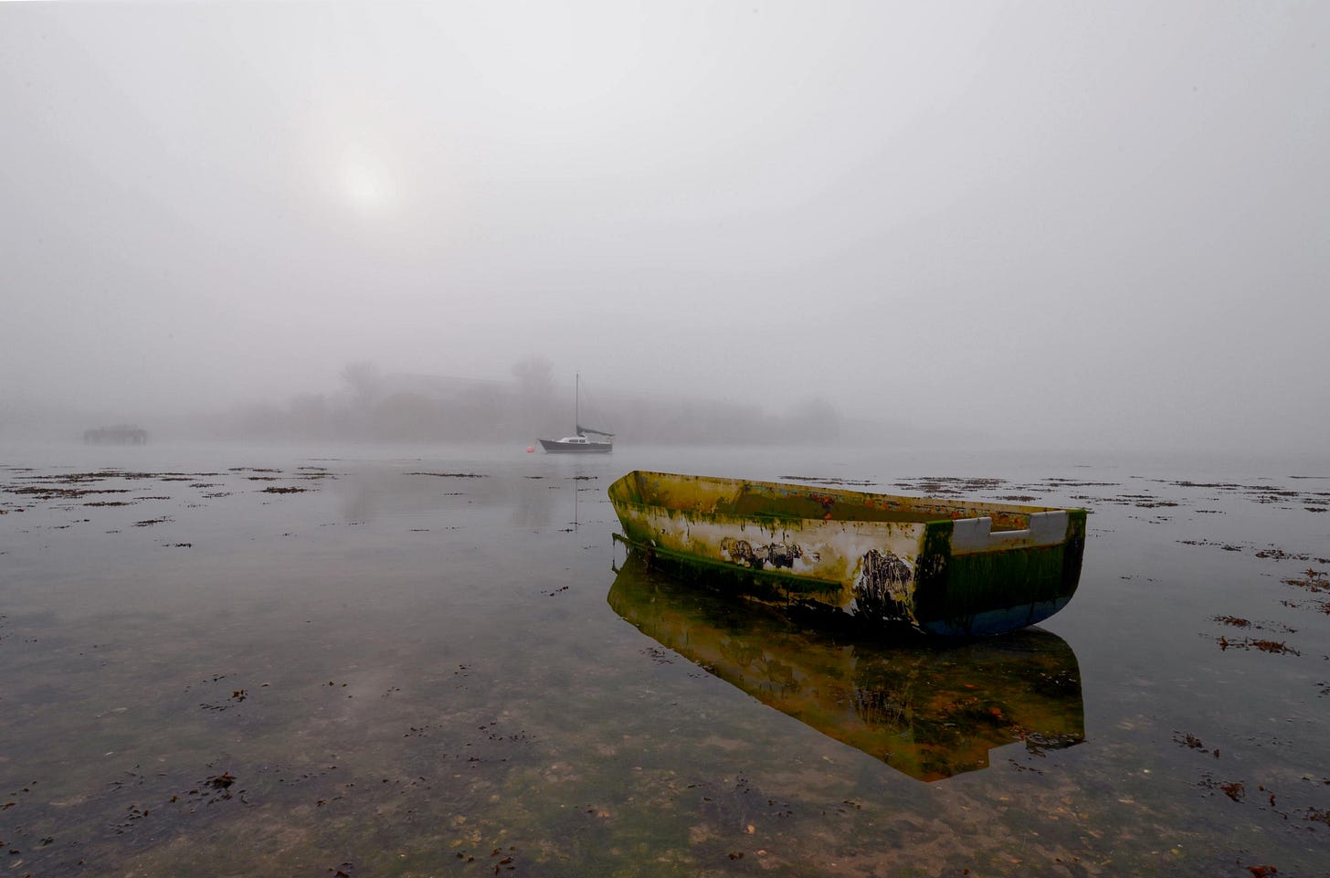A quiet, atmospheric photograph of an abandoned boat resting on still water, partially obscured by fog, capturing a sense of isolation, stillness and quiet reflection.