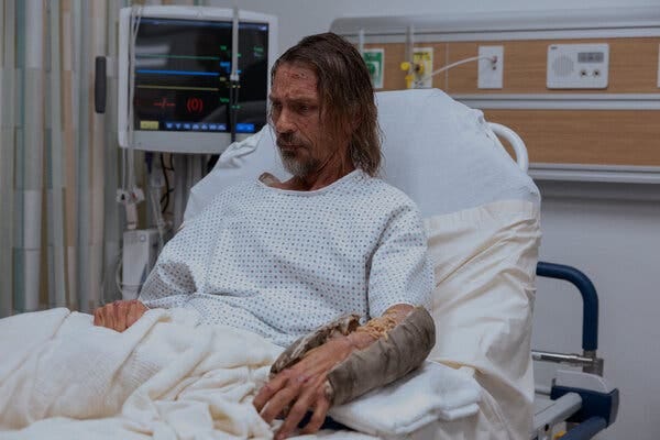 A bedraggled man in a hospital gown lies in a hospital bed with his dirty cast cut open to reveal maggots on his arm.