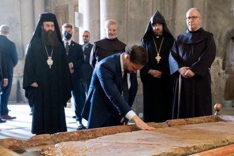 Several Orthodox clergy members in black robes and tall hats with crosses stand around a man in a dark suit kneeling and touching a large flat stone slab on the floor of a stone church interior with columns and arches in the background. Several Orthodox clergy members in black robes and tall hats with crosses stand around a man in a dark suit kneeling and touching a large flat stone slab on the floor of a stone church interior with columns and arches in the background.