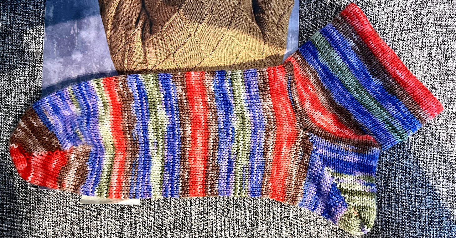 A knitted sock in stripes of blue, red, green, and brown, shown from above on a grey upholstered surface. A knitted sock in stripes of blue, red, green, and brown, shown from above on a grey upholstered surface.