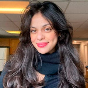 Mulher jovem olhando para a câmera, com a cabeça levemente inclinada. Tem cabelos longos, escuros e volumosos, com ondas leves, caindo sobre os ombros. A pele é clara a morena clara, e os olhos parecem castanhos. Veste uma blusa preta de gola alta, usa batom em tom rosa e está sorrindo. No nariz, há um piercing em forma de argola no septo, e nas orelhas, pequenos brincos. com um tecido que parece macio e ajustado ao corpo.