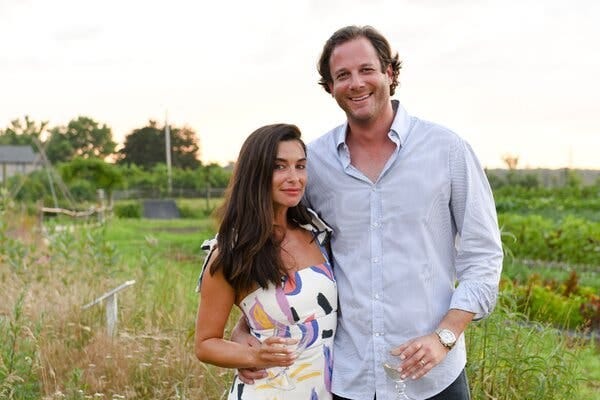 Candice Miller, in a white summer dress, poses for a portrait with her husband, Brandon Miller, in a light blue shirt. Both are holding wine glasses. Candice Miller, in a white summer dress, poses for a portrait with her husband, Brandon Miller, in a light blue shirt. Both are holding wine glasses.