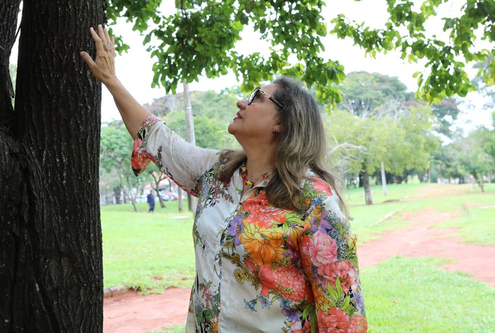 Pessoa com cabelos longos e óculos escuros, vestindo blusa floral colorida, toca o tronco de uma árvore em um parque arborizado com gramado verde e caminho de terra ao fundo.