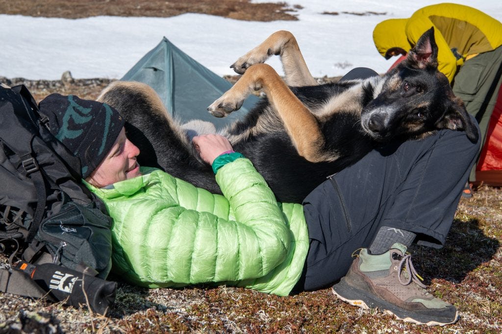 A woman in a bright green down jacket and beanie lies back on the tundra, smiling, with a black and tan dog sprawled belly-up across her legs, a tent and snow patches behind.