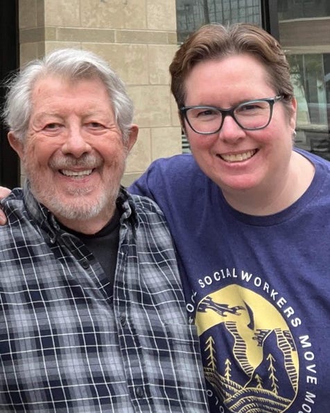 A warm photo of Frank Falk and me from April 2022. Frank, white-haired and bearded, wears a blue and black plaid shirt and smiles broadly. I'm next to him in dark-framed glasses and a navy t-shirt that reads "Colorado School Social Workers Move Mountains," with my arm around his shoulders. Both of us are smiling at the camera.