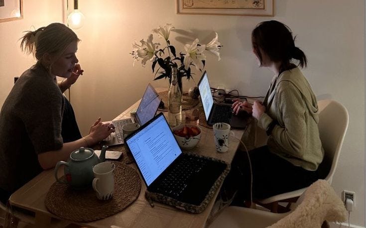 This may contain: two women sitting at a table with laptops on their lap tops and coffee mugs in front of them This may contain: two women sitting at a table with laptops on their lap tops and coffee mugs in front of them