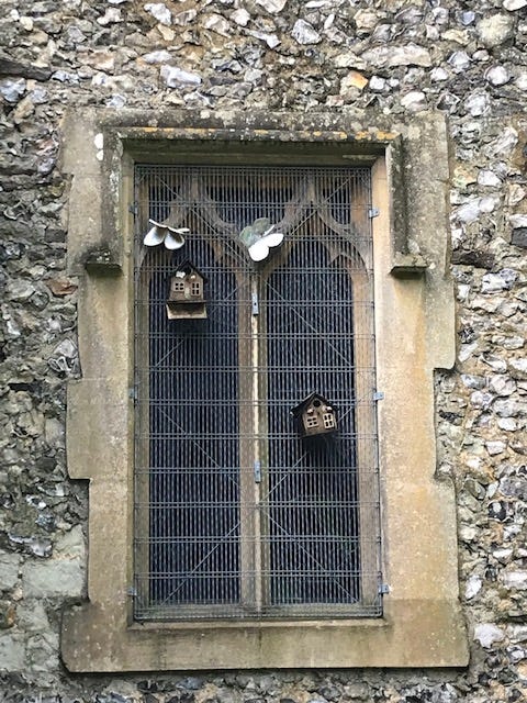 Stony East Wall of the Chaldon Church, highlighting a window, protected by a grille. There are bird feeders and butterflies made of what might be metal, attached to the window grille