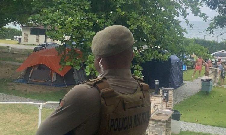 A uniformed military police officer with a beige beret and tactical vest stands facing a grassy campsite with tents and trees. In the background, nude and clothed individuals walk near camping equipment and buildings under an overcast sky.
