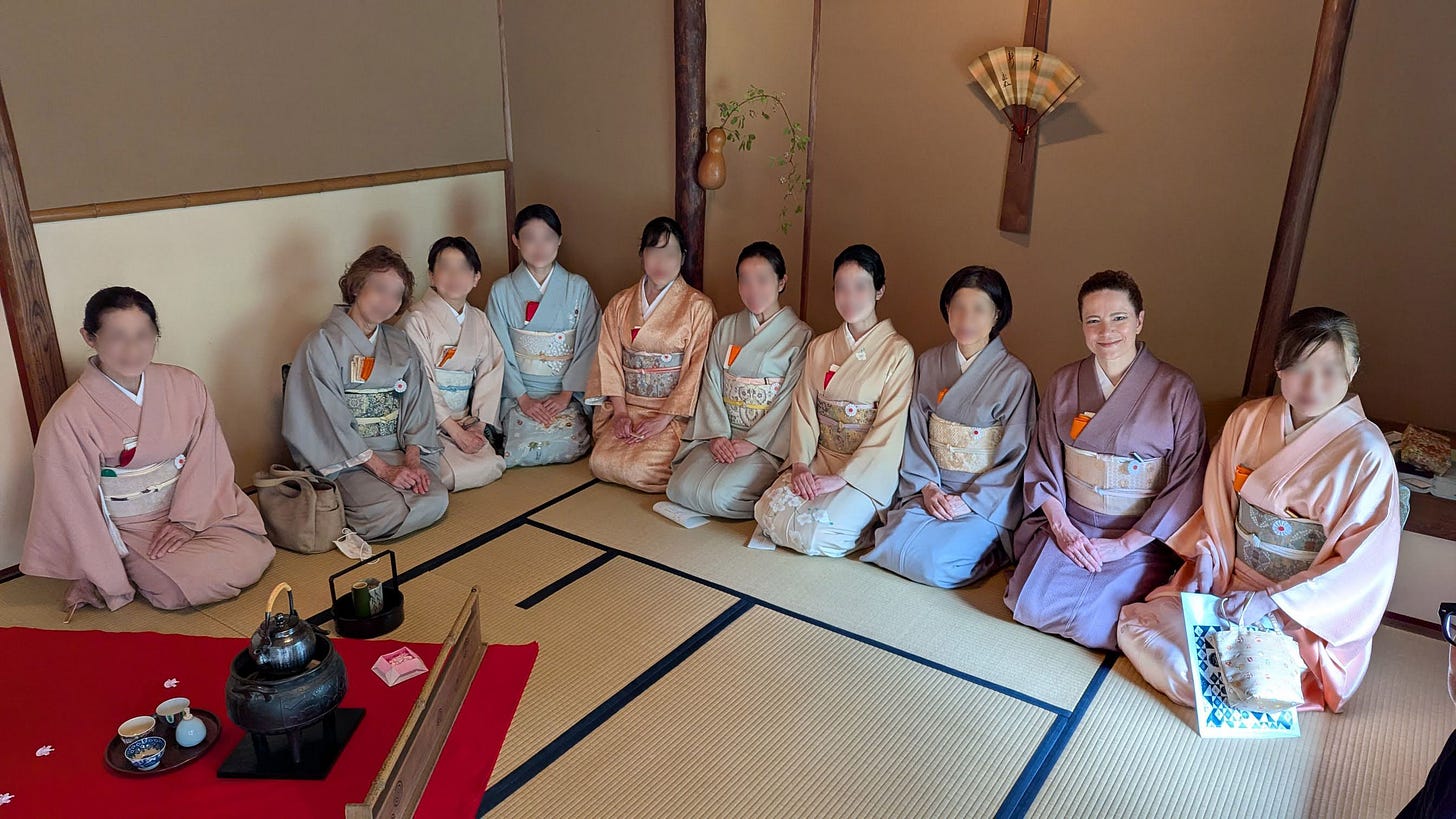 Women in pastel kimono sit on the floor in a tatami room where a small tea kettle and other utensils are displayed on a red floor mat. In the alcove, a flower arragement hangs on a wooden post and there's a large fan displayed on the wall.