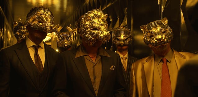 Men in suits wear ornate gold animal masks, standing in a dimly lit room with reflective walls, creating a mysterious and opulent atmosphere.