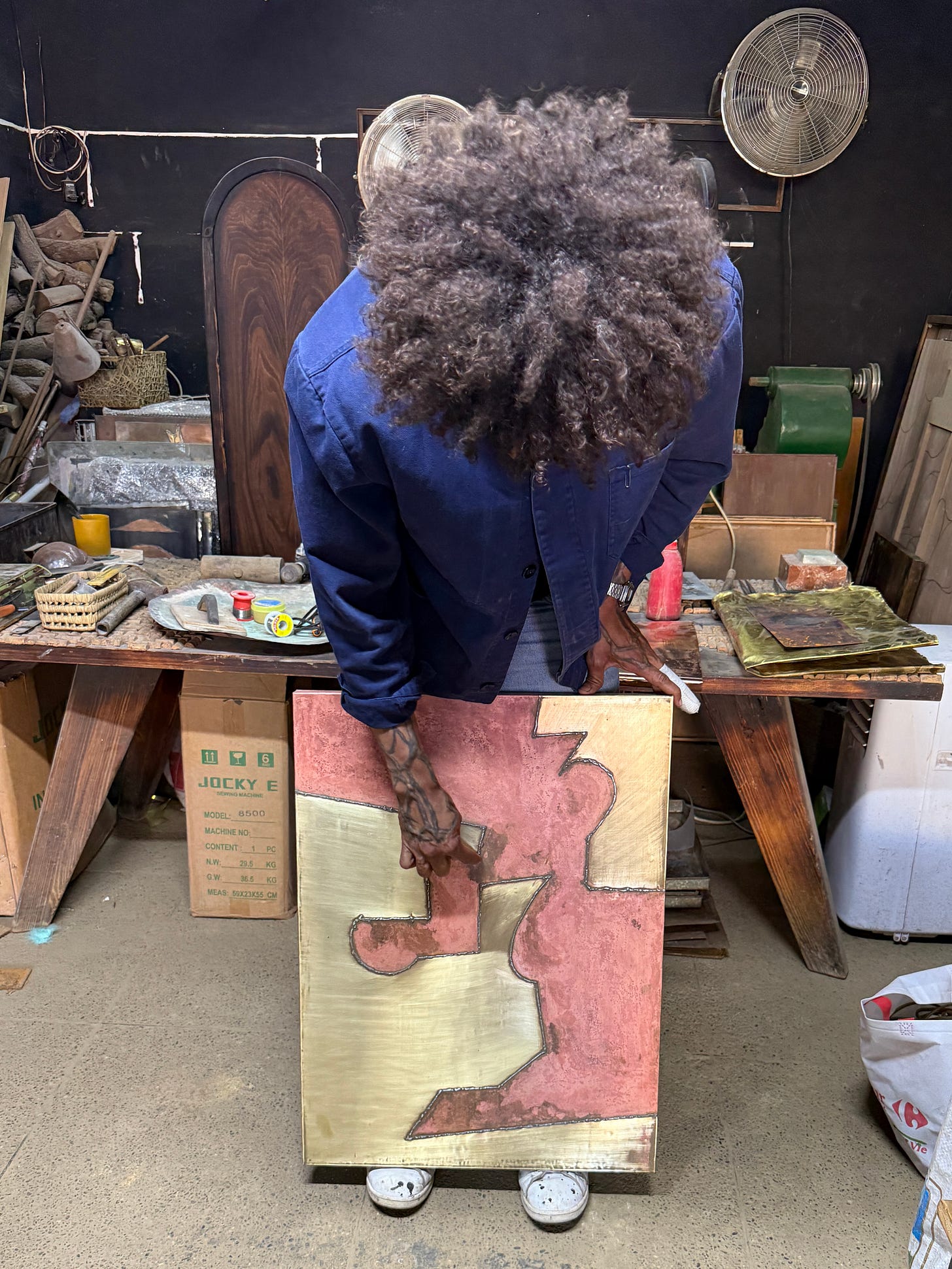 Artist Abdessamia Bargamane seen from above in his workshop in Marrakech, bending over a metal artwork placed against his legs. The studio is cluttered with tools, metal sheets, pigments, and worktables, emphasizing a hands-on, craft-based artistic process. Artist Abdessamia Bargamane seen from above in his workshop in Marrakech, bending over a metal artwork placed against his legs. The studio is cluttered with tools, metal sheets, pigments, and worktables, emphasizing a hands-on, craft-based artistic process.