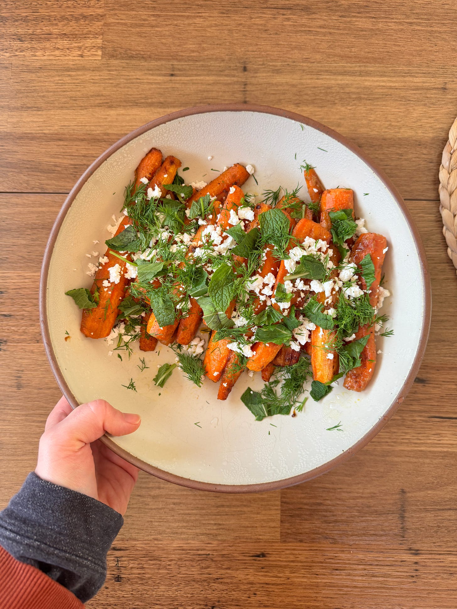 A hand comes into frame holding a ceramic serving bowl with roasted carrots topped with crumbled feta, mint, and dill.