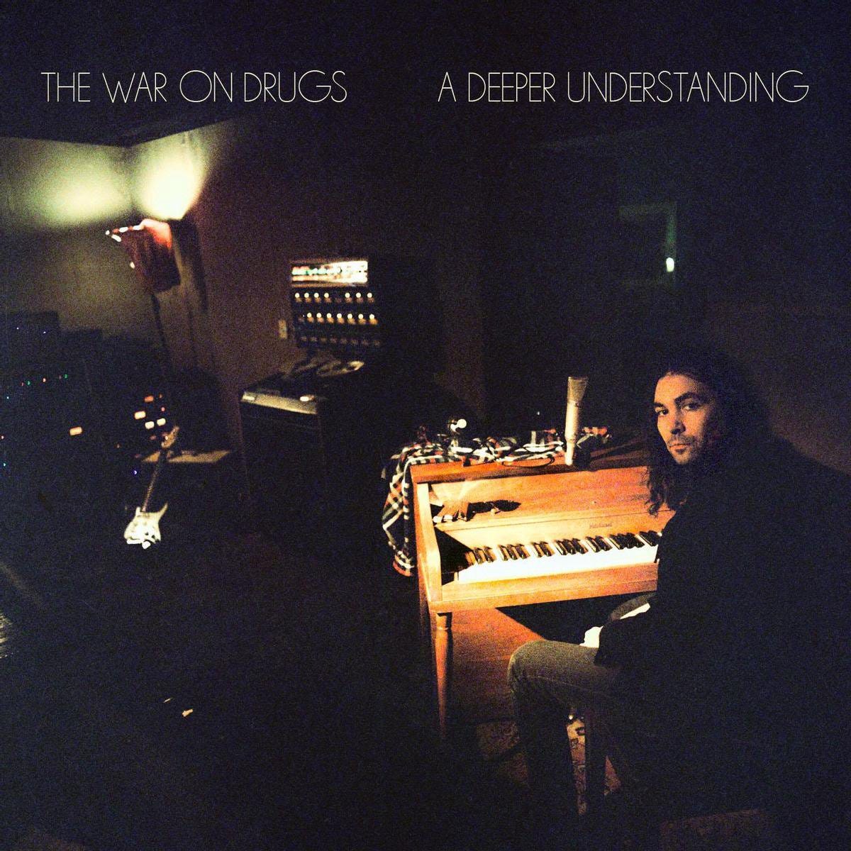 War On Drugs frontman Adam Granduciel sits in a dark studio room, in front of an upright piano. The piano is glowing, and Granduciel is turned towards the viewer, as though taking a pause from his work. In the shadows of the background, are various pieces of audio equipment: lights from consoles glitter in the dark, and the face of an electric guitar can be seen as well.