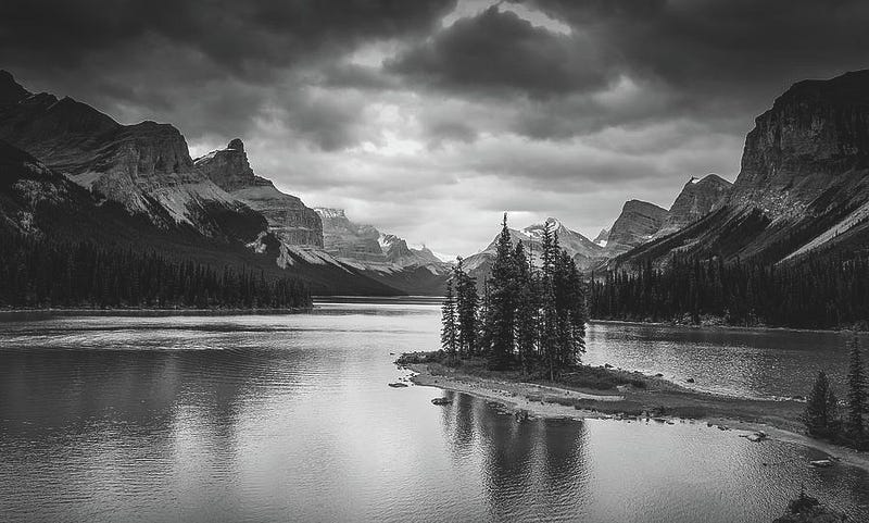 A black and white photograph of Spirit Island at Maligne Lake, showing trees on a small island framed by towering mountains and calm reflective water.