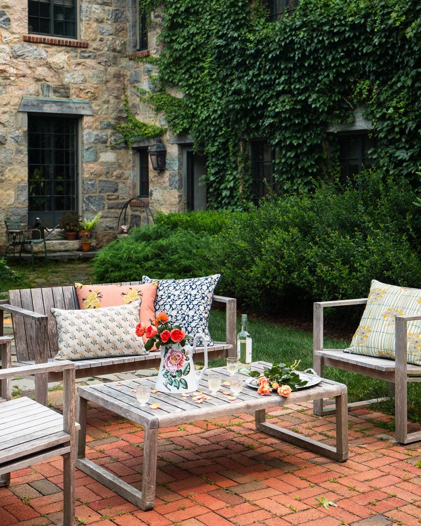Outdoor patio furniture set in a stone courtyard with ivy-covered walls, featuring a weathered wooden coffee table, matching armchairs, and patterned throw pillows with a vase of red flowers on a brick patio.