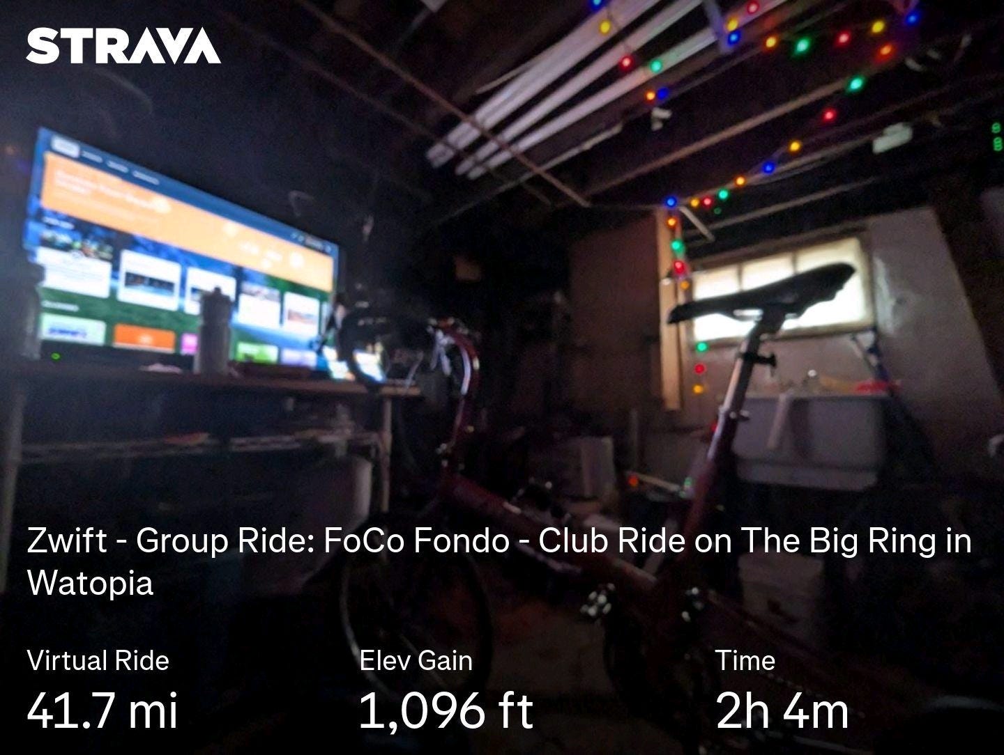 Photo of a dark basement. To the right is a small window letting some outside light in. Nearby, hanging from the rafters are some holiday lights. To the left, on a table, is a flat screen tv with the Zwift cycling app on it. In the dim foreground is a bicycle on a stationary trainer.