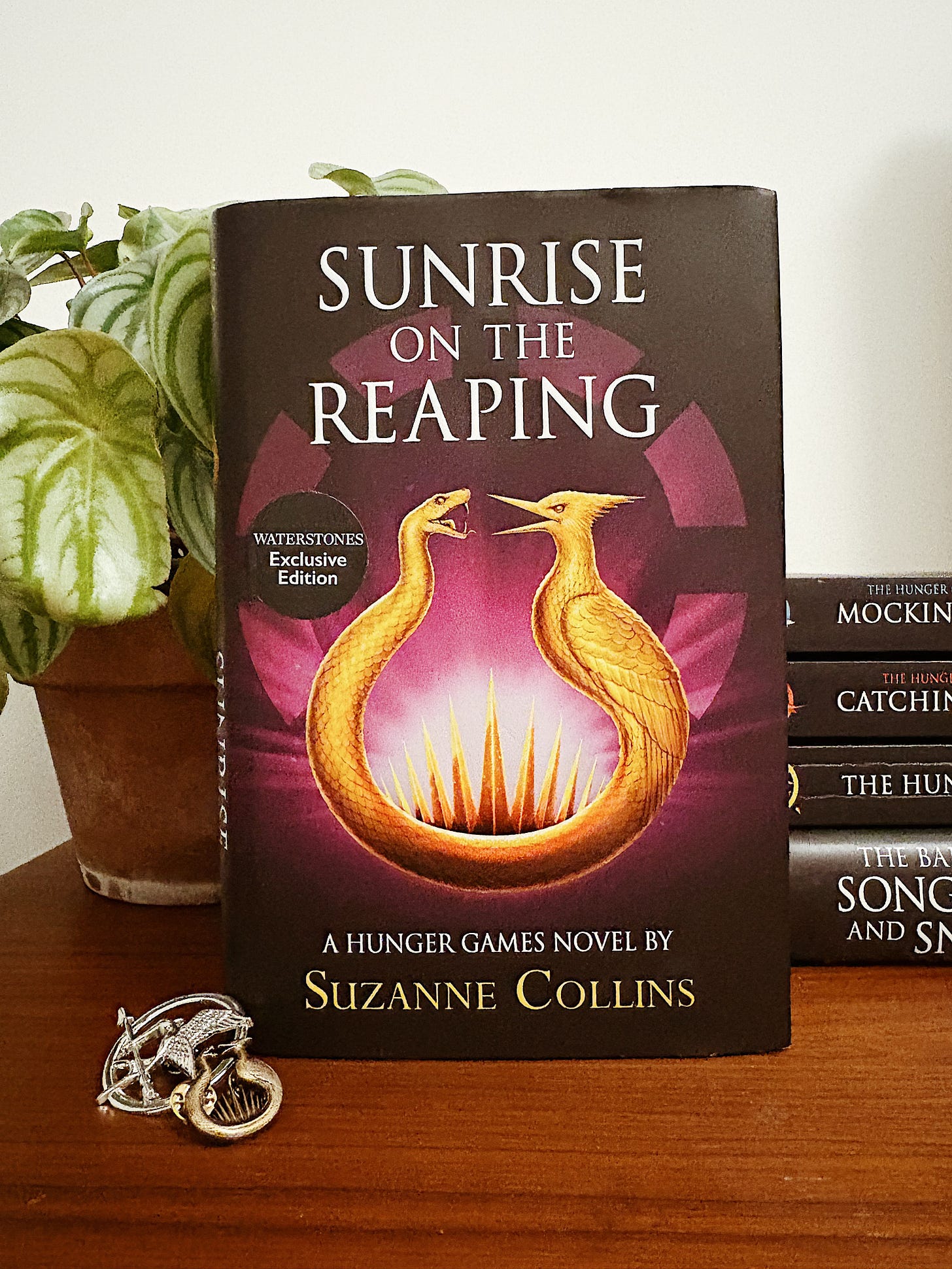 A copy of Sunrise on the Reaping placed on a wooden table. To the left, a plant and two mockingjay-themed pins. To the right, the rest of the Hunger Games books piled in chronological order.
