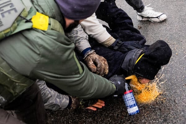 A man in green gear sprays a can of chemical irritant in the face of a man who is being held down by other people. 