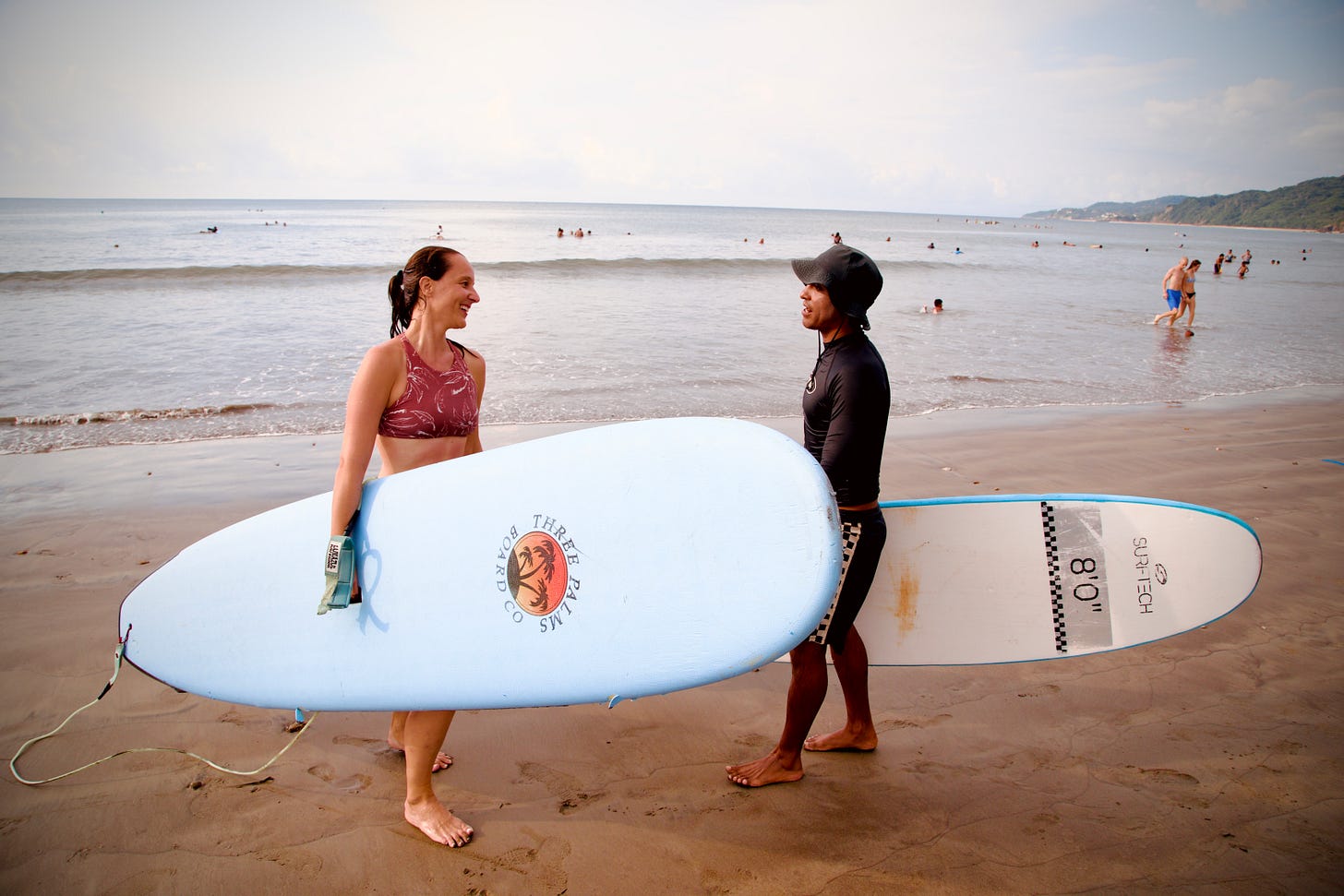 Two people standing on beach holding large white surfboard together - woman in red swimsuit and man in black wetsuit and cap, with ocean waves and other beachgoers in background. Photograph by Georges Yazbek