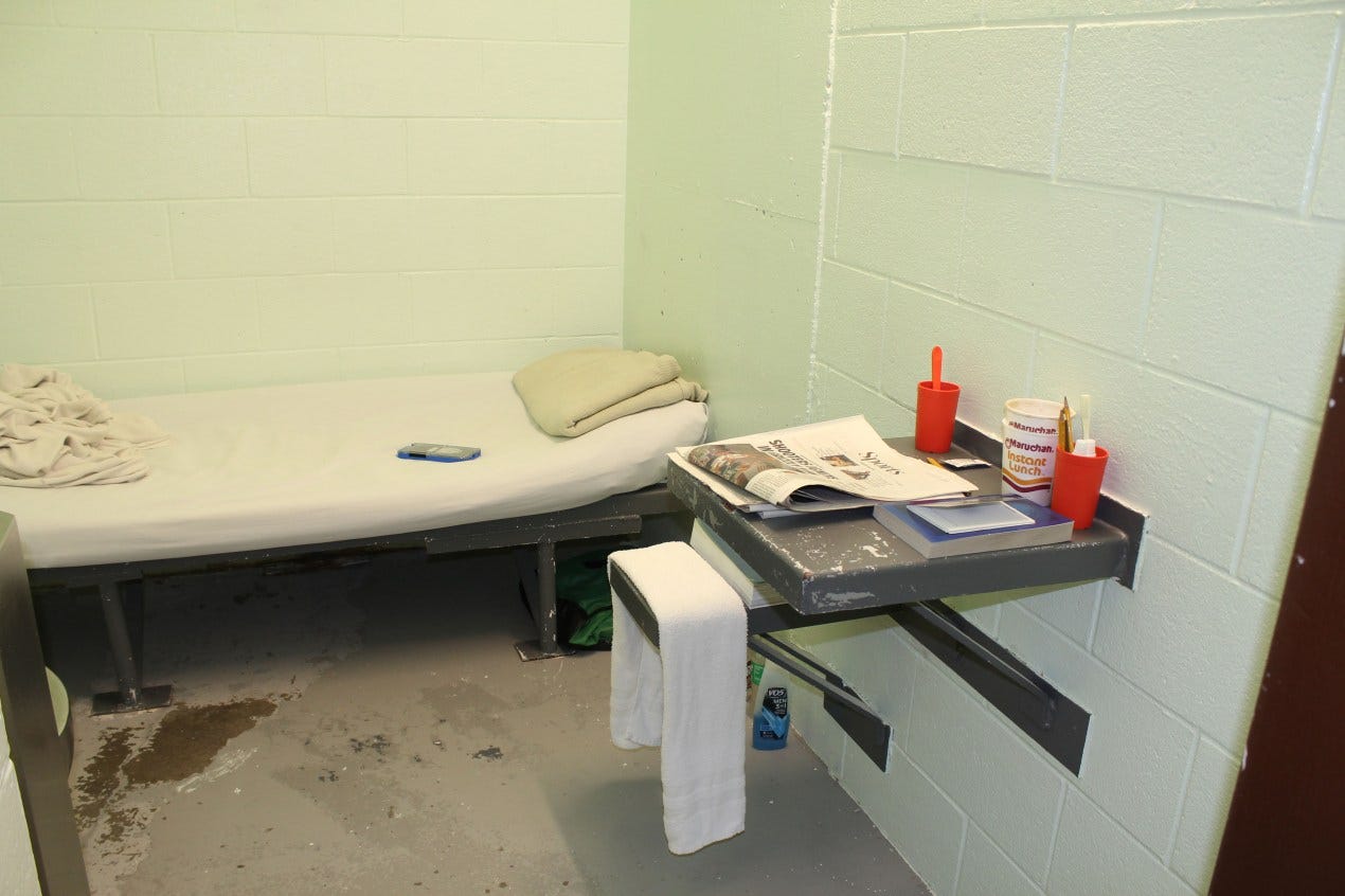 A small prison cell. There is a narrow bed agains the wall with dilapidated sheets, and a metal table connected to the wall. A small prison cell. There is a narrow bed agains the wall with dilapidated sheets, and a metal table connected to the wall.