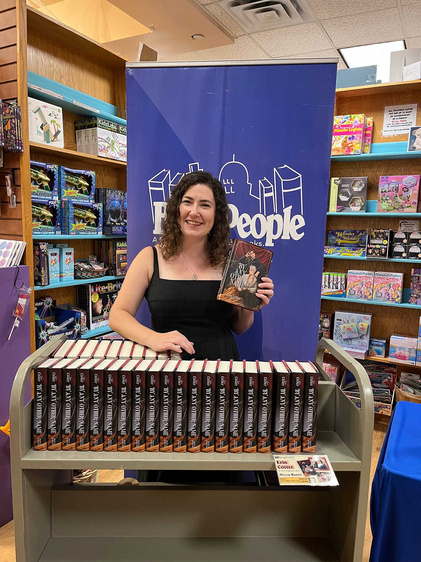 a white woman in a black jumpsuit holds up a copy of her book above a library cart filled with more copies of her book. 
