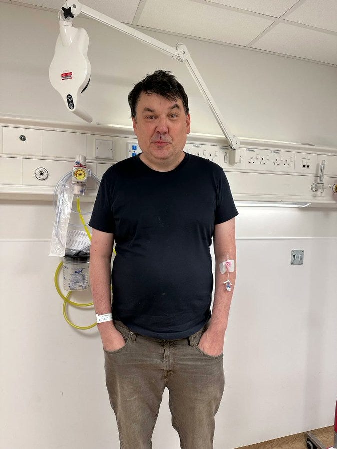 Graham Linehan standing in a hospital room. He wears a black t-shirt and brown pants, with a hospital bracelet on his wrist and medical tape on his arm. Medical equipment, including a light fixture and wall-mounted devices, is visible in the background.