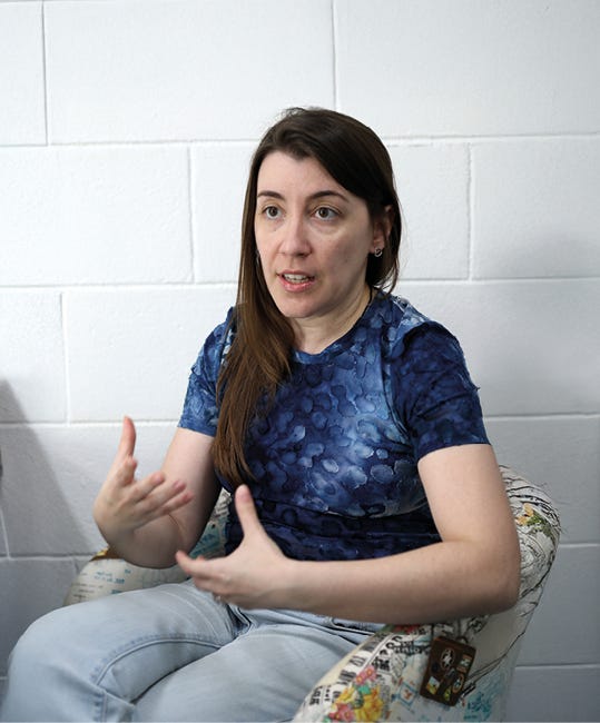 Pessoa sentada em cadeira estampada, usando camiseta azul-marinho com padrão de círculos, calça jeans clara, com cabelo castanho comprido, gesticula com ambas as mãos enquanto fala, posicionada de frente para a câmera contra parede branca de tijolos aparentes. Pessoa sentada em cadeira estampada, usando camiseta azul-marinho com padrão de círculos, calça jeans clara, com cabelo castanho comprido, gesticula com ambas as mãos enquanto fala, posicionada de frente para a câmera contra parede branca de tijolos aparentes.