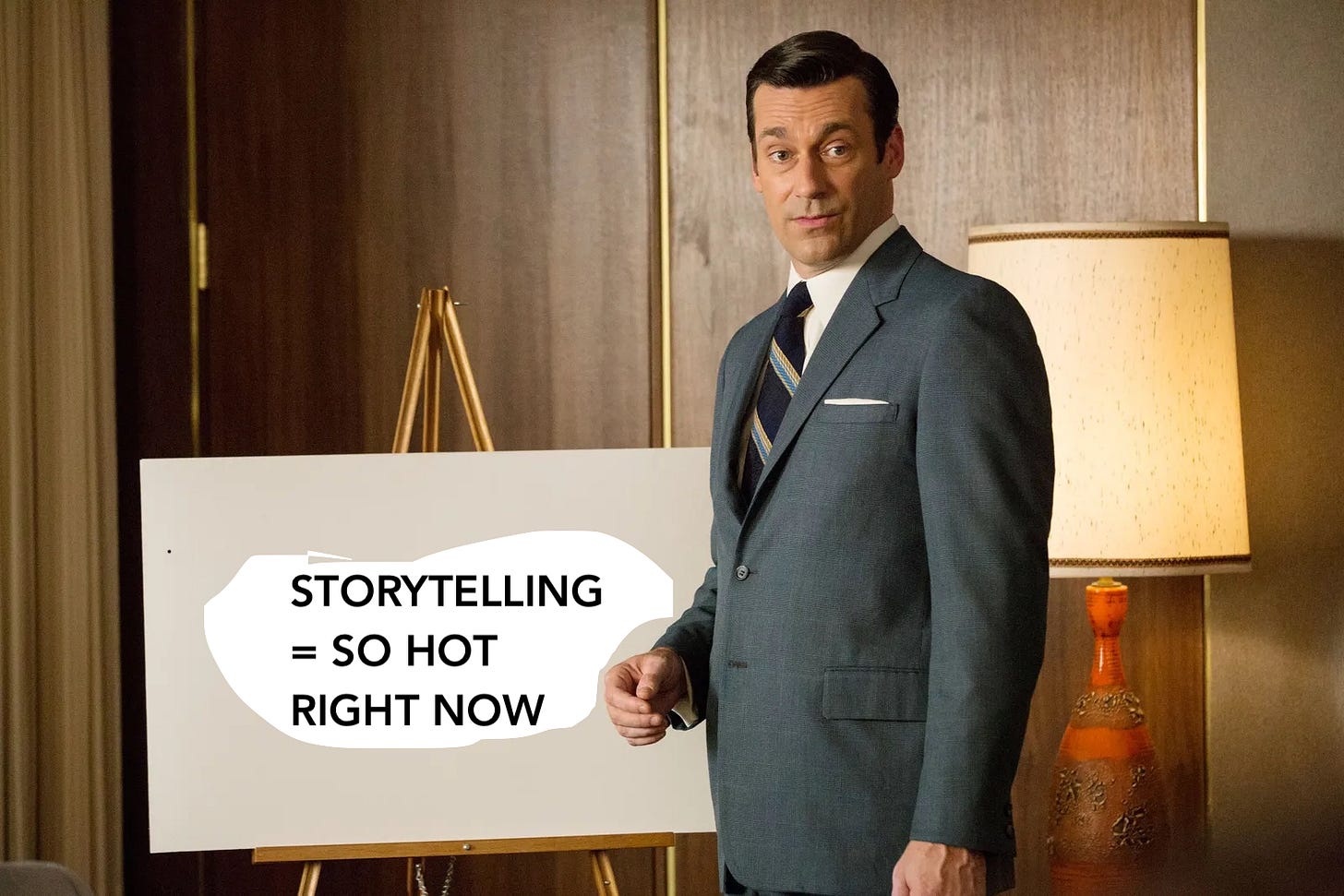 A 1960's ad executive named Don Draper, in a gray suit, looks a little confused in a wood paneled room. He's standing in front of an easel. I've edited the easel to say: "Storytelling = So Hot Right Now."