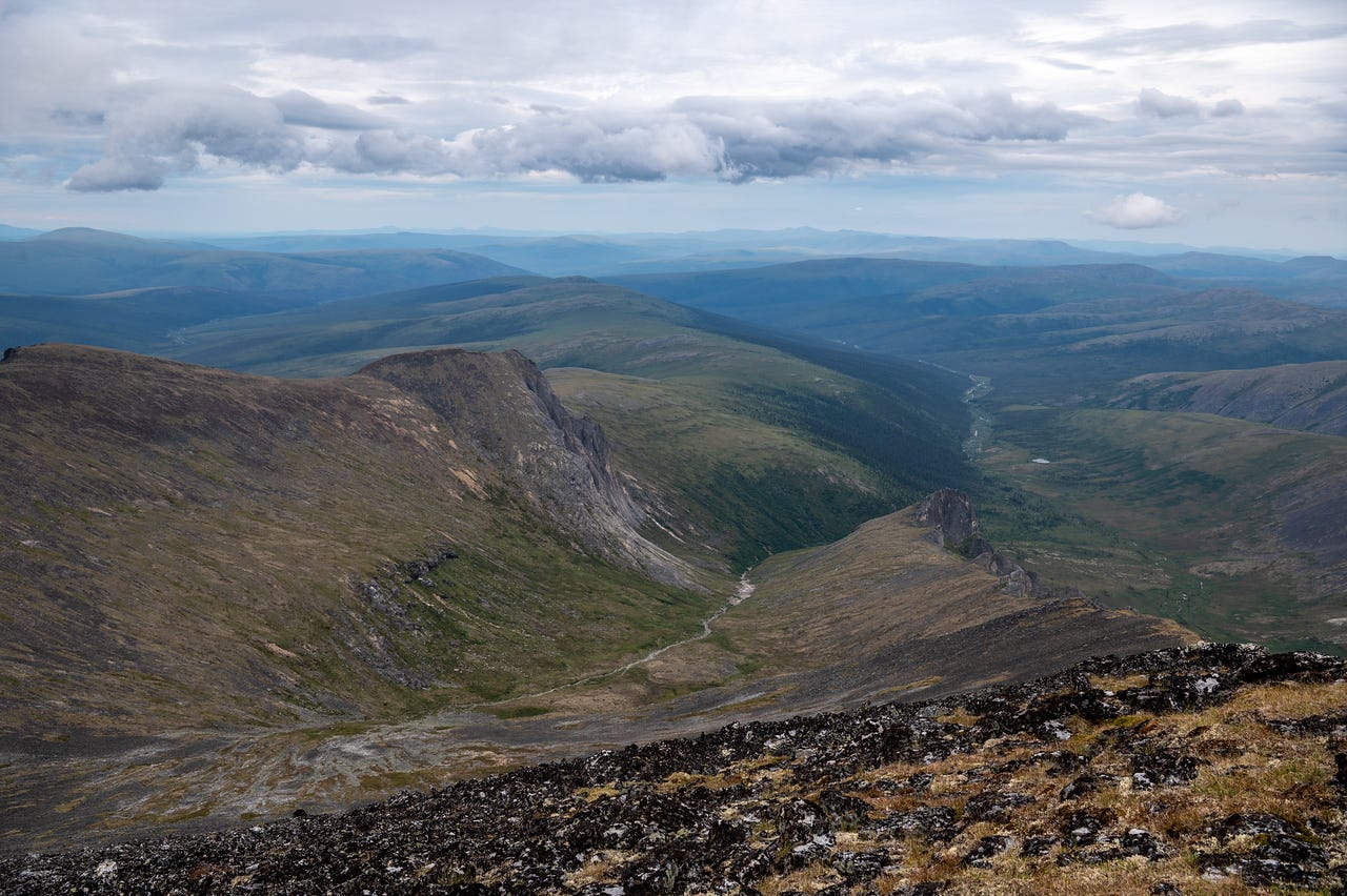 A deep, U-shaped valley drops away below the summit, green tundra floor threaded by a braided stream running out toward spruce and distant blue ranges, steep rubble slopes rising on both sides under broken cloud.