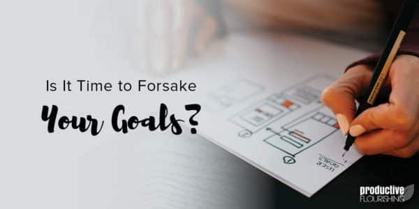 A tan hand writes on a paper on top of a dark table. Text Overlay: Is It Time To Forsake Your Goals? A tan hand writes on a paper on top of a dark table. Text Overlay: Is It Time To Forsake Your Goals?