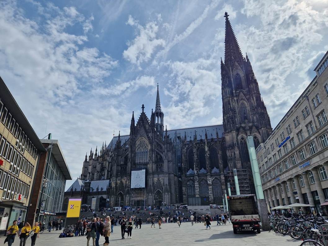 Side view of the catheral in Cologne with a square in front of it and stairs leading up to it. Houses on either side. One is a hotel. 