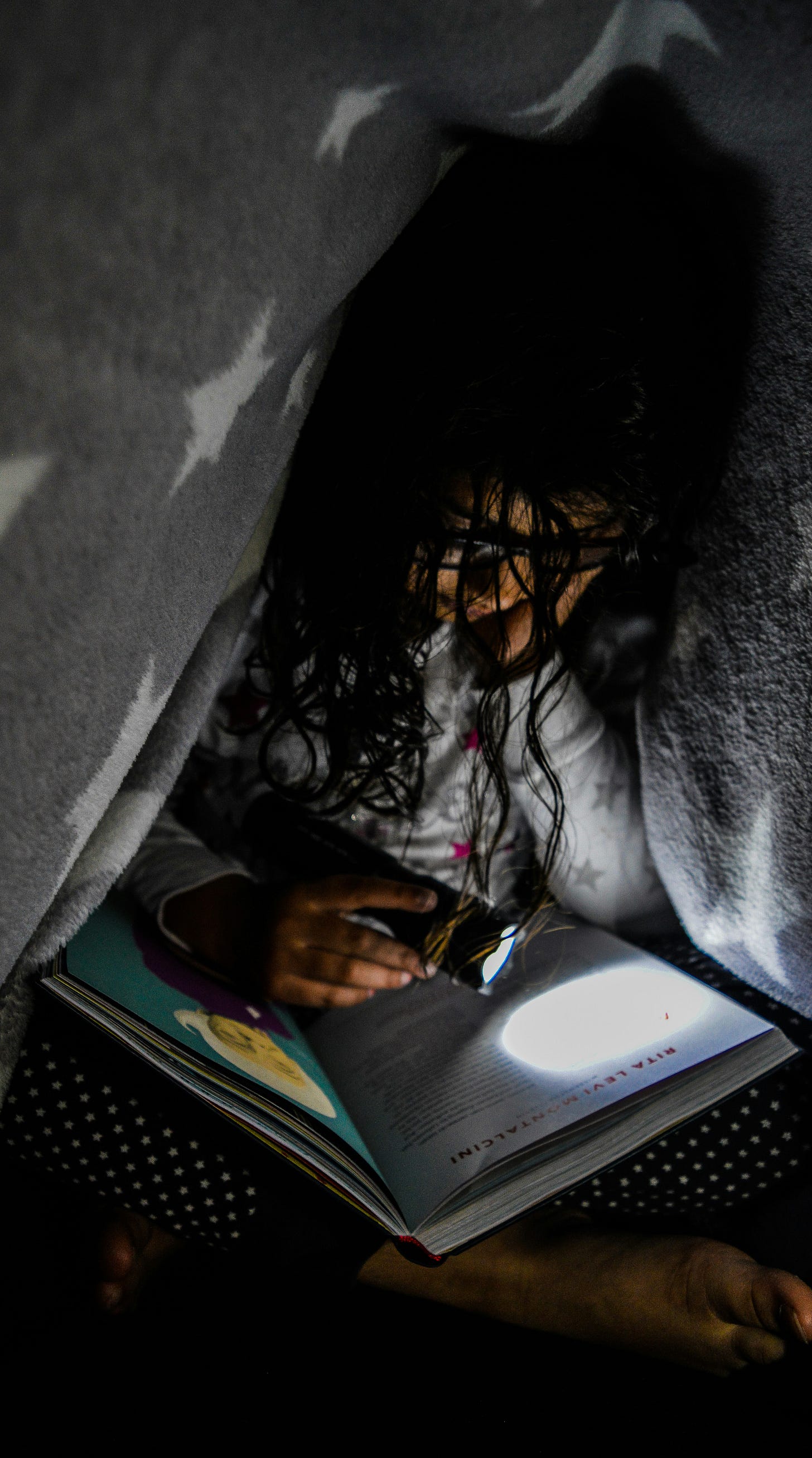 Child reading a book inside a blanket fort using a small flashlight, suggesting a dark room due to a broken lamp or unavailable light source.