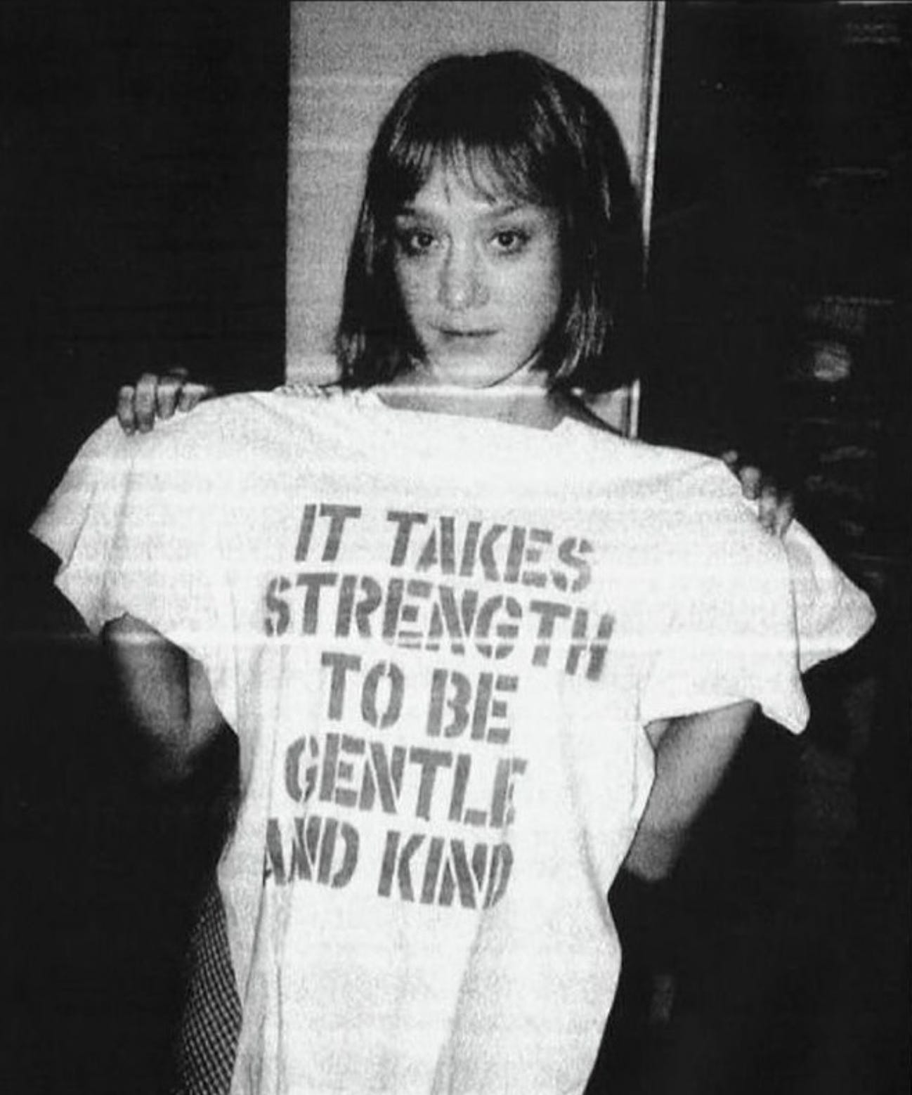 Photo of Chloë Sevigny holding up a white tee-shirt that reads "IT TAKES STRENGTH TO BE GENTLE AND KIND."