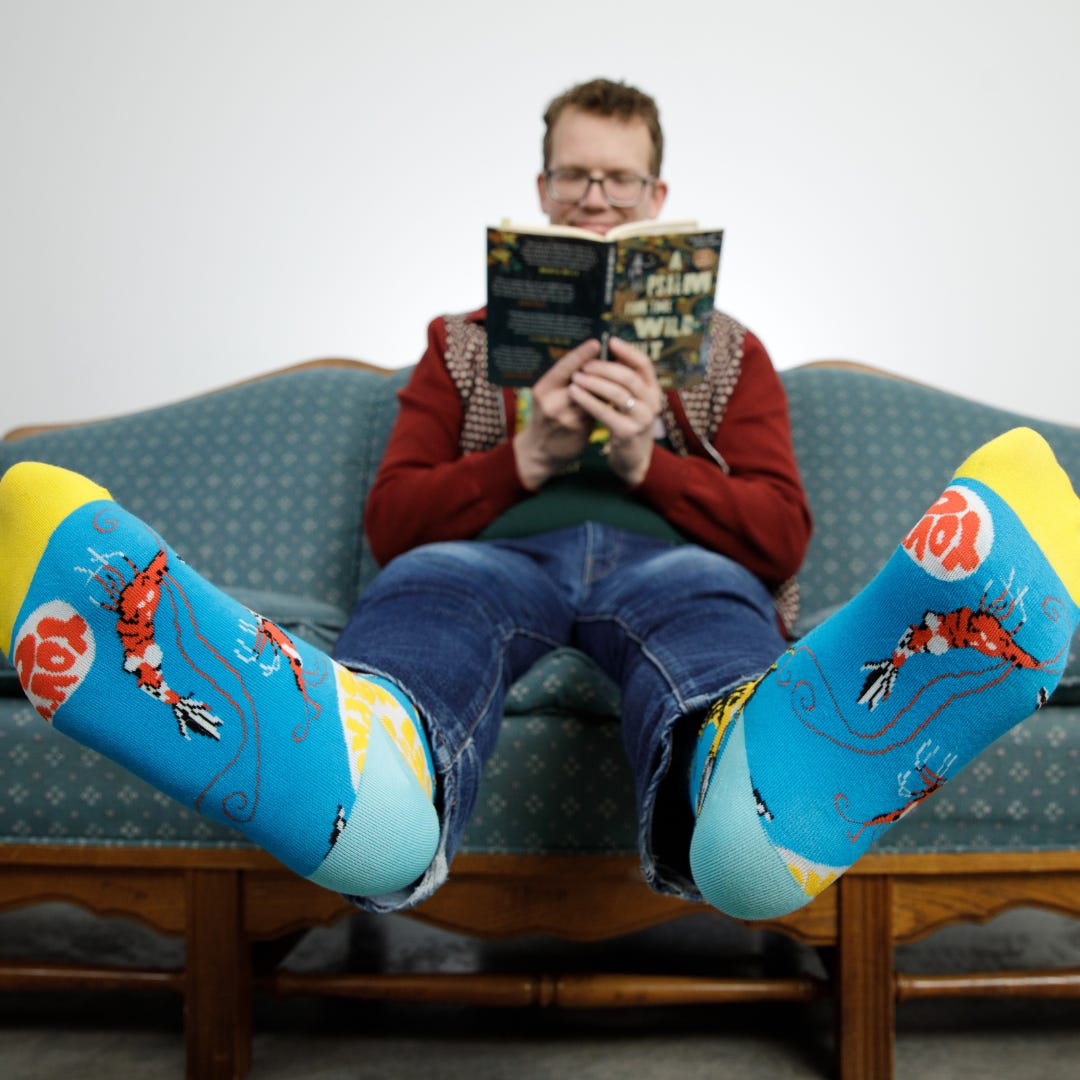 A promotional shot of a man wearing shrimp-themed socks while sitting on a couch reading a book. His feet are in the foreground, in sharp focus, while he is in the background in soft focus.
