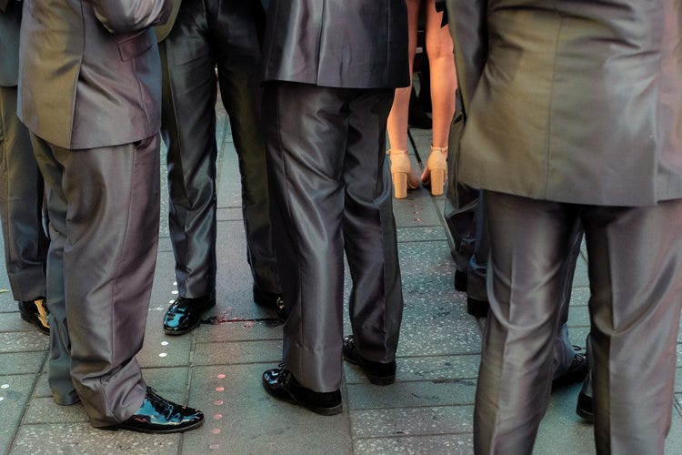 Foto colorida de Craig Whitehead mostrando um monte de pernas masculinas vestindo roupas sociais cinzas e, no meio delas, duas pernas femininas trajando meias-calças rosadas.