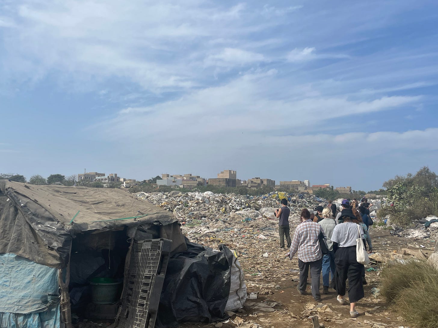 A group of people walks through a large landfill under a partly cloudy sky. The ground is littered with trash, and makeshift structures covered with tarps can be seen in the foreground. In the background, low-rise buildings are visible beyond the heaps of waste.