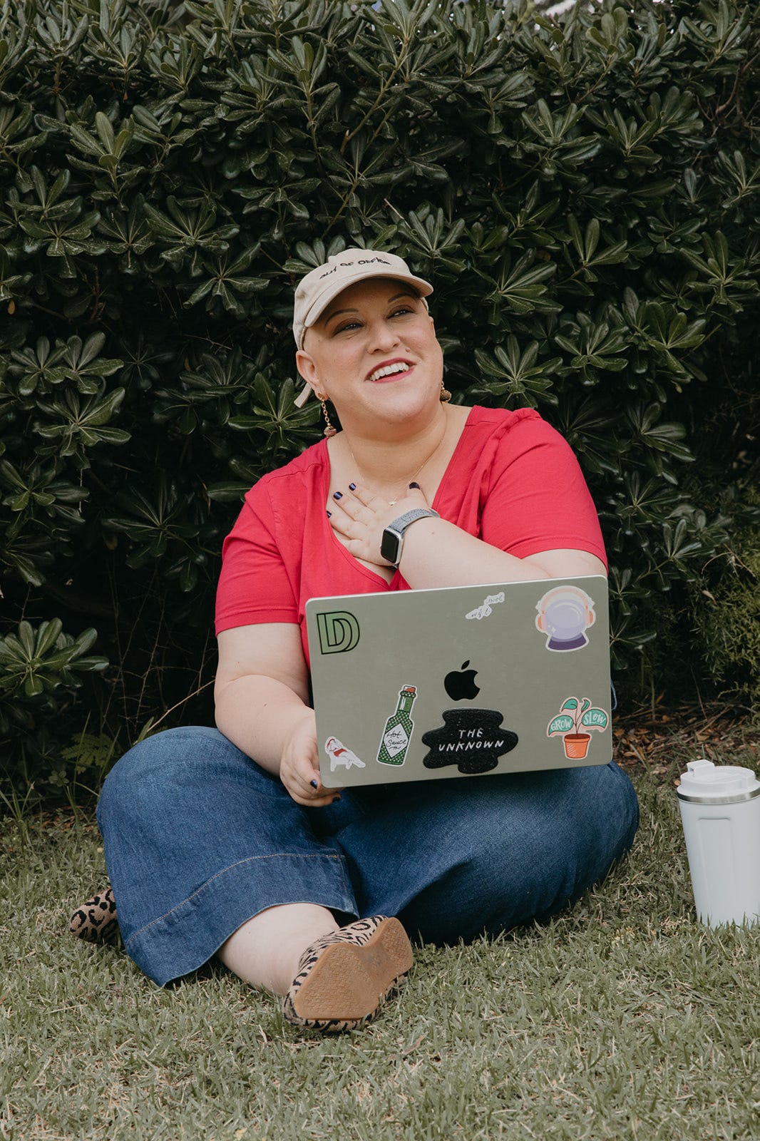 Chedva Vivi Ludmir sitting on a patch of grass with trees in the background wearing a pink shirt and a baseball cap, laughing and holding a hand to her chest with a laptop in her lap