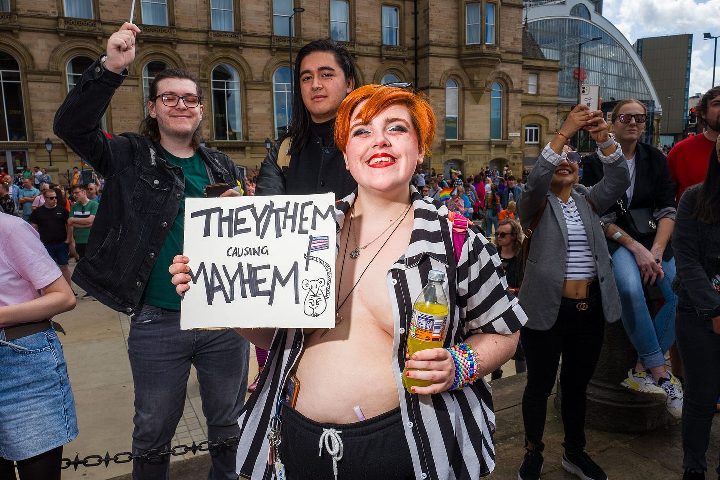 A person in a black and white striped open shirt with their chest partly exposed, smiles and holds a sign saying "They/them causing mayhem"