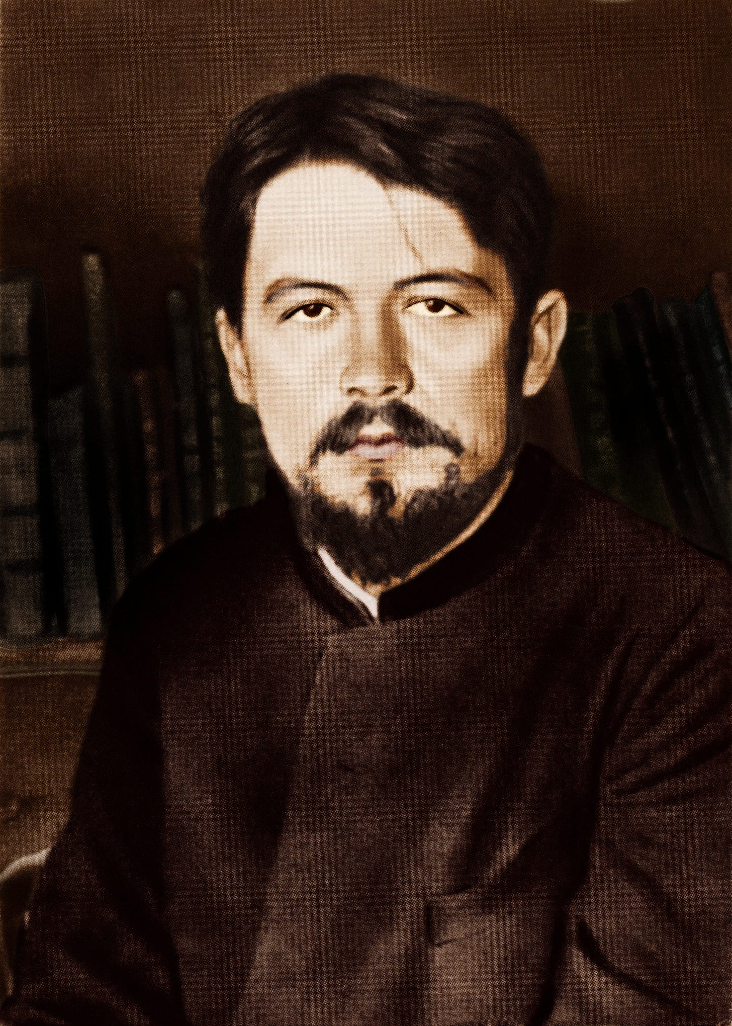 Sepia-toned photograph of the writer Anton Chekhov, with moustache and pointed beard, seated indoors in a dark high-collared coat, with books blurred in the background. Chekhov's expression is sombre.