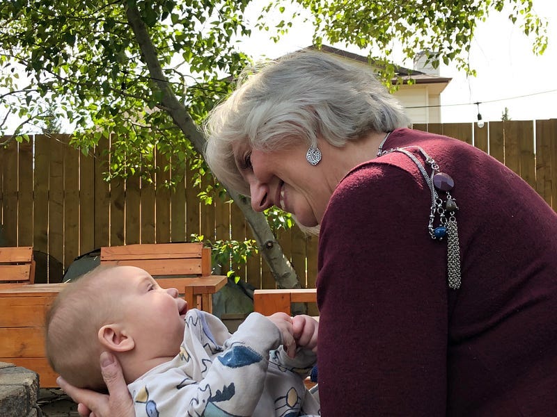 A baby boy looks up at his Grandma as she looks back at him with adoration. She has thrown her long chain necklace over her shoulder so he won’t get tangled in it.