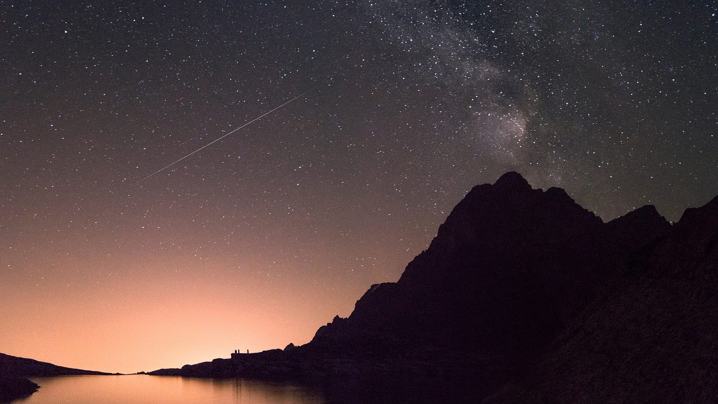 Shooting Star streaks across the night sky, light illuminates the horizon revealing the edges of a mountain and a body of water