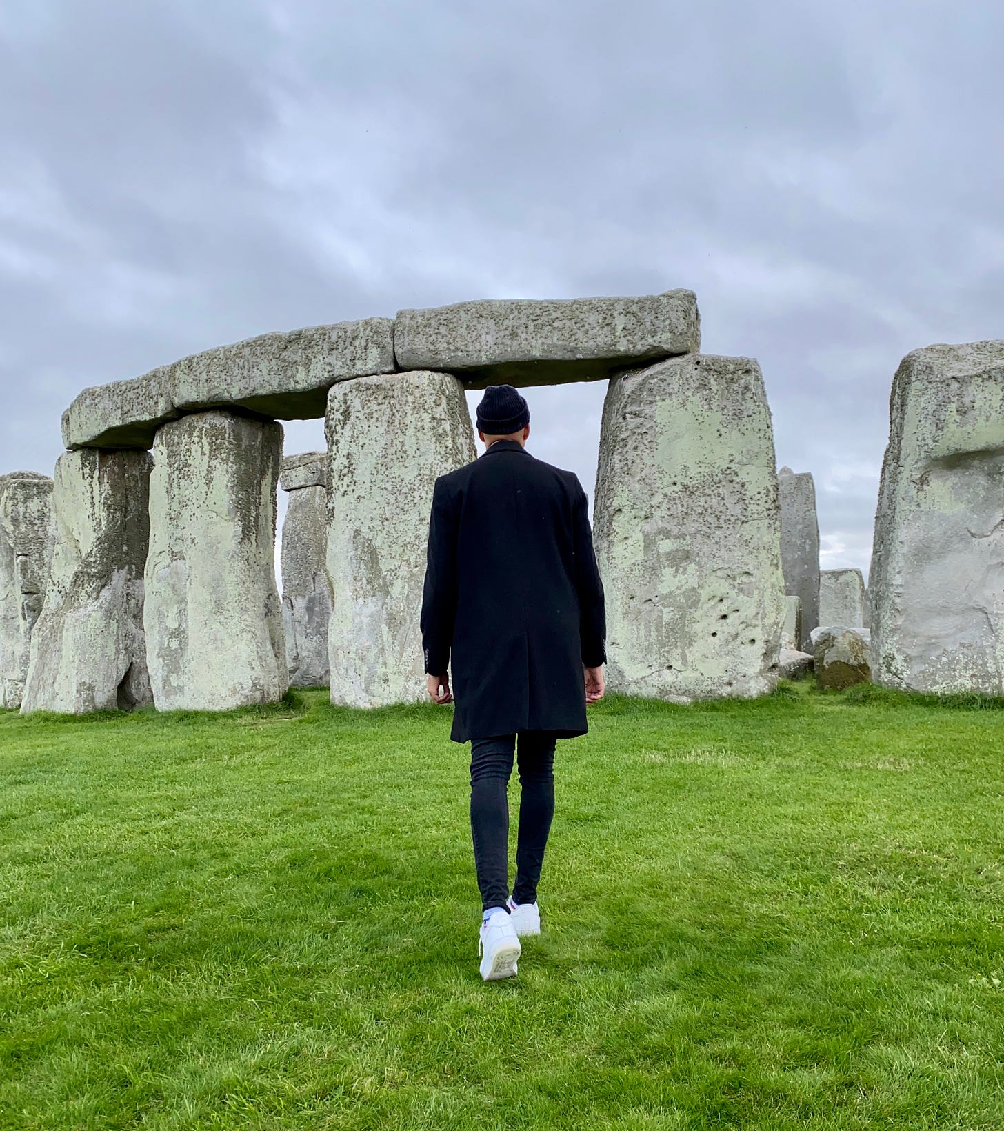 Ethan Ward in a black coat and beanie walks toward the stone archway at Stonehenge on an overcast day. The grass is green and the site is empty.
