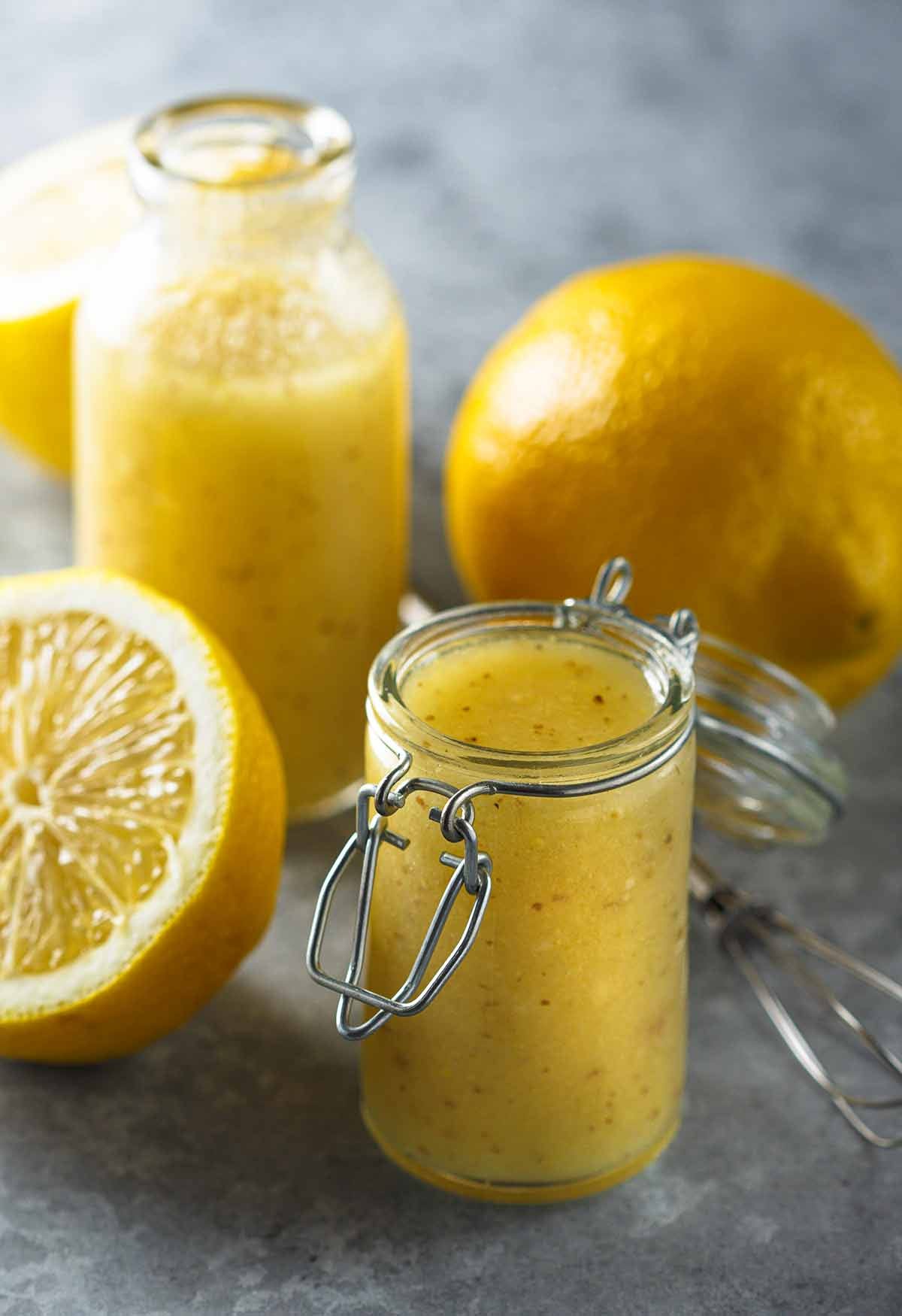A clear glass jar filled with bright, emulsified lemon vinaigrette featuring visible herbs and cracked black pepper.