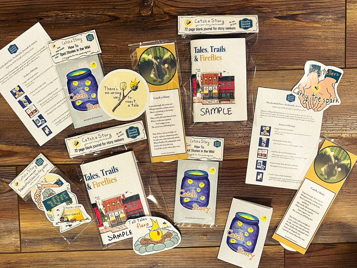 A collection of Catch a Story product samples laid out on a wooden surface. Items include zines, bookmarks, stickers, and product information sheets. Designs feature illustrated fireflies in jars, campfires, hands holding a glowing spark, and a small-town street scene titled Tales, Trails & Fireflies. Some items are labeled “SAMPLE” and packaged in clear sleeves, and there are circular stickers with quotes like “There’s no wrong way to roast a tale” and “Stories shine when shared.”