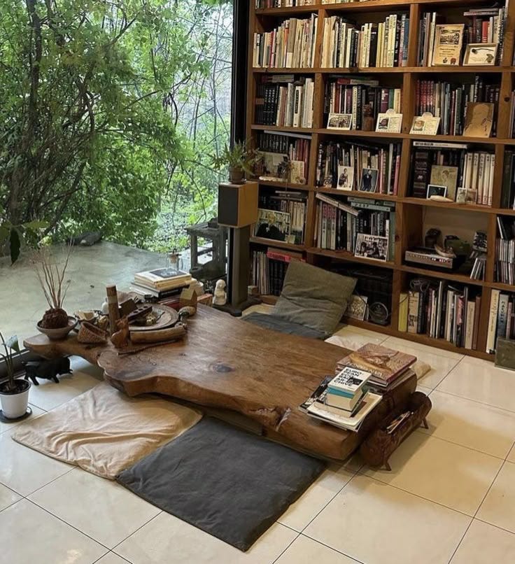 This may contain: a living room filled with lots of books on top of a wooden table next to a window