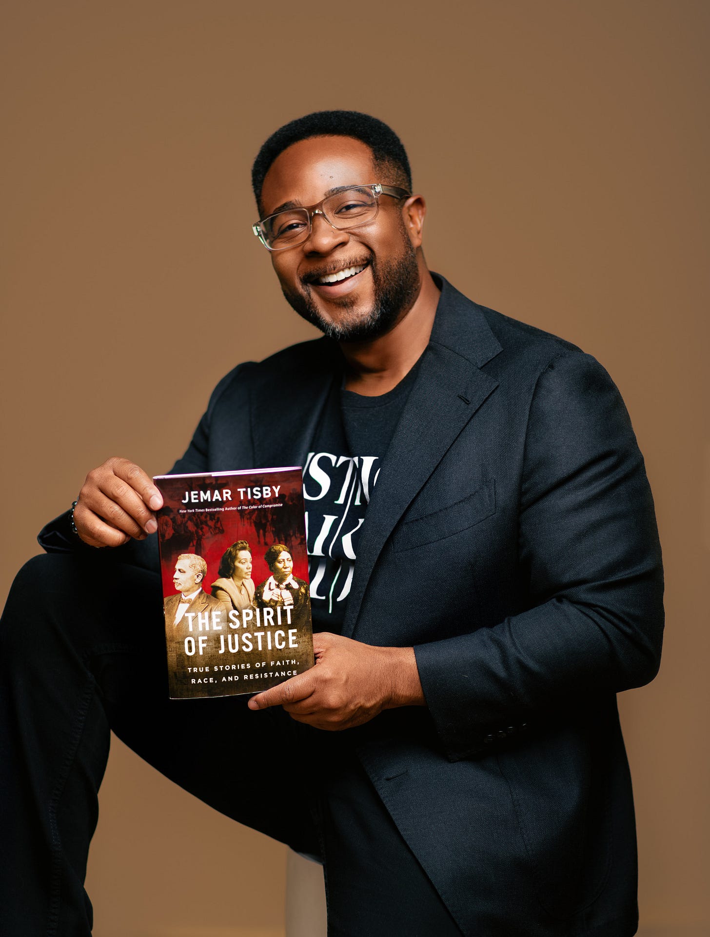 Jemar Tisby in all-black w/ Justice Takes Sides t-shirt smiling holding a copy of The Spirit of Justice