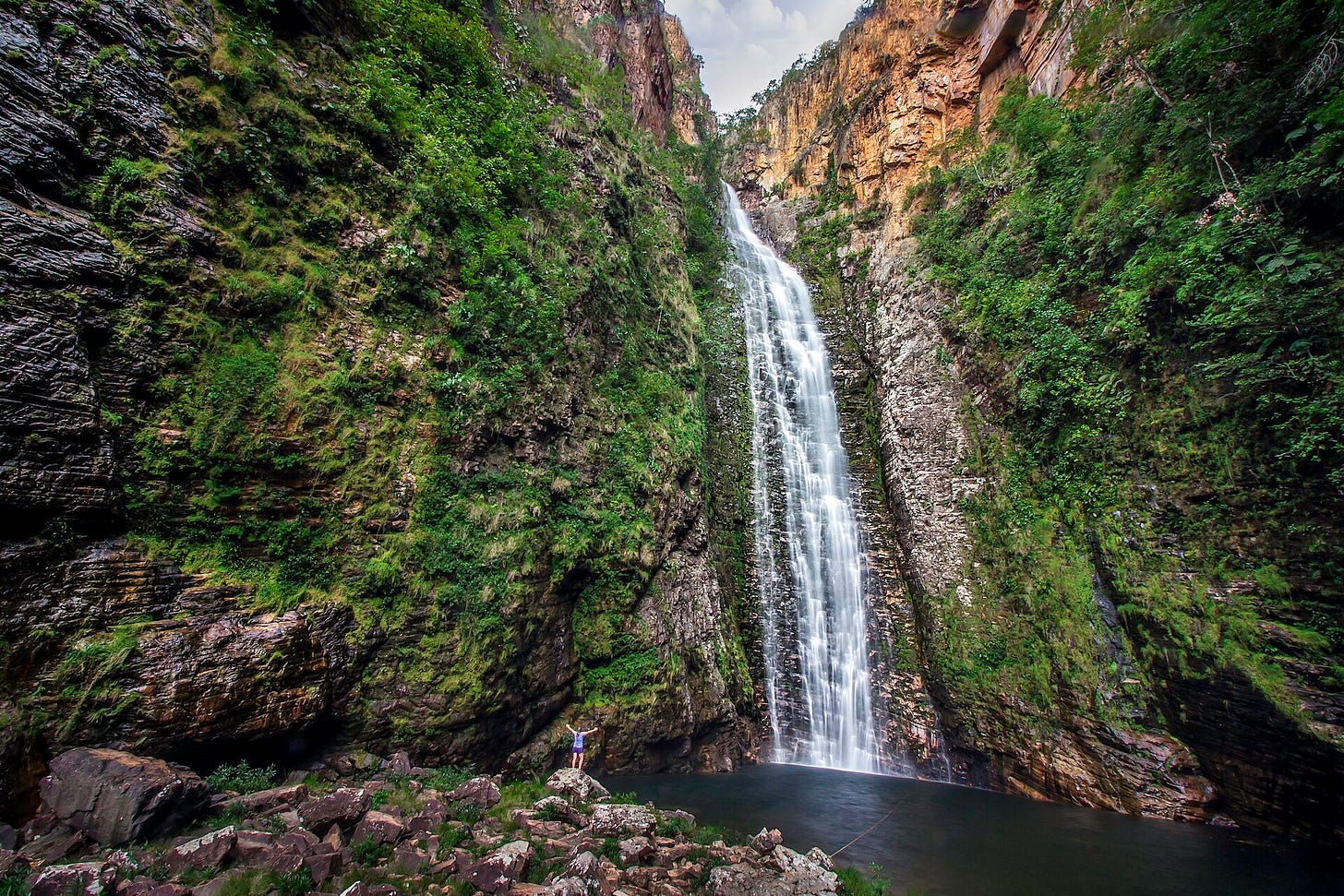 A lone figure stands with arms raised at the base of the Cachoeira do Segredo, a 115-meter waterfall dropping through a narrow canyon of moss-covered rock walls in the Chapada dos Veadeiros, Goiás, Brazil. A lone figure stands with arms raised at the base of the Cachoeira do Segredo, a 115-meter waterfall dropping through a narrow canyon of moss-covered rock walls in the Chapada dos Veadeiros, Goiás, Brazil.